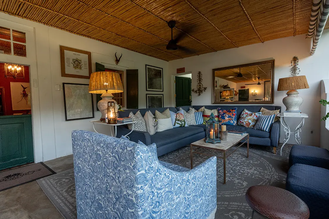 Perfect Hideaways property for sale: Cozy living room with blue-patterned chairs, a large L-shaped sofa with colorful cushions, and a rustic coffee table. Warm lighting and art adorn the walls. Daniel’ Kraal Farm.
