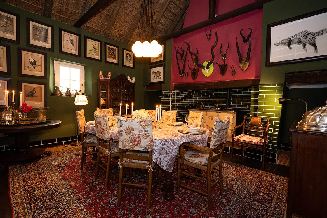 Perfect Hideaways property for sale: Cozy dining room with a rustic vibe, featuring a large table set for dinner with floral chairs. Walls adorned with framed bird art and antelope horns. Warm lighting from a chandelier and candles. Daniel’ Kraal Farm.
