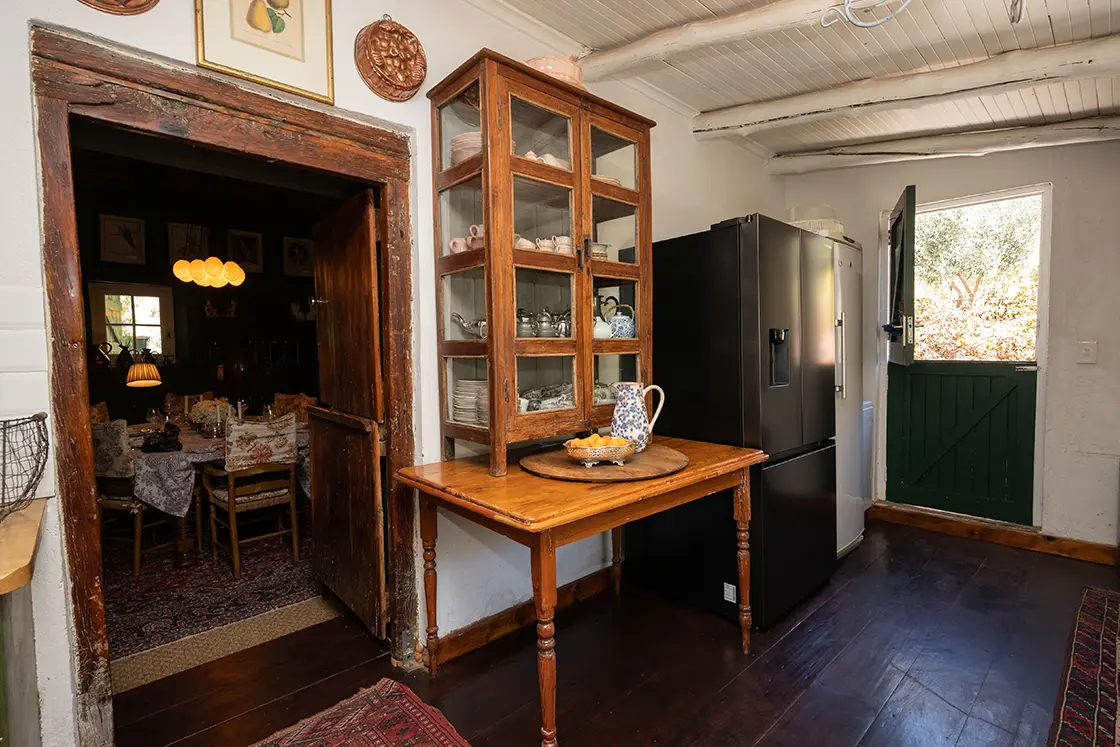 Perfect Hideaways property for sale: Cozy kitchen with vintage decor; wooden table with china jug, glass cabinet, black fridge, open door to sunny garden, and rustic dining room visible. Daniel’ Kraal Farm.