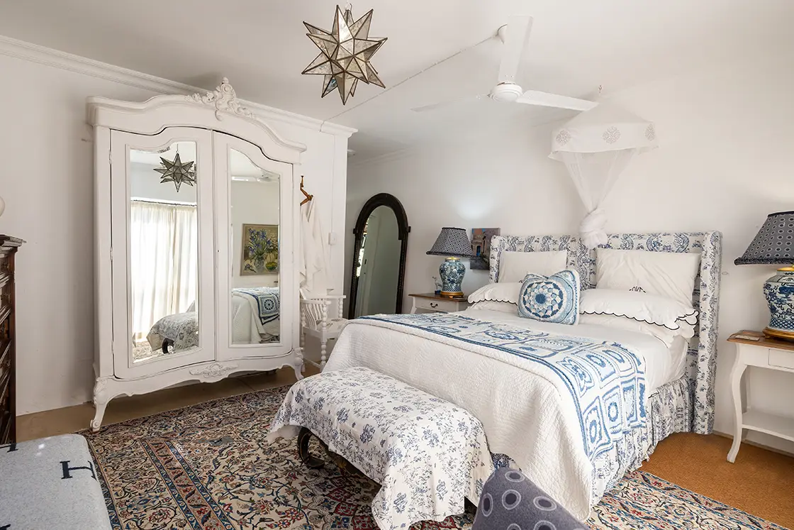 Perfect Hideaways property for sale: Elegant bedroom with a white and blue theme. Features a patterned bedspread, vintage armoire with mirrors, star-shaped pendant light, and an ornate rug. Daniel’ Kraal Farm.