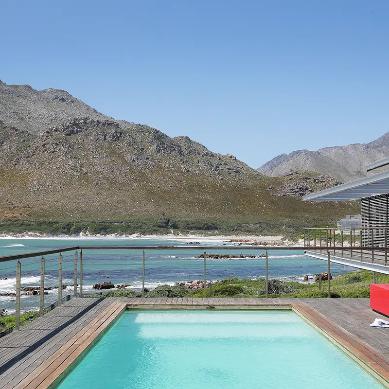 Perfect Hideaways property for sale: Die Punt, Rooi Els. Modern coastal villa with a pool overlooking the ocean. Wooden deck features a red bench, while mountains provide a serene backdrop under a clear blue sky.