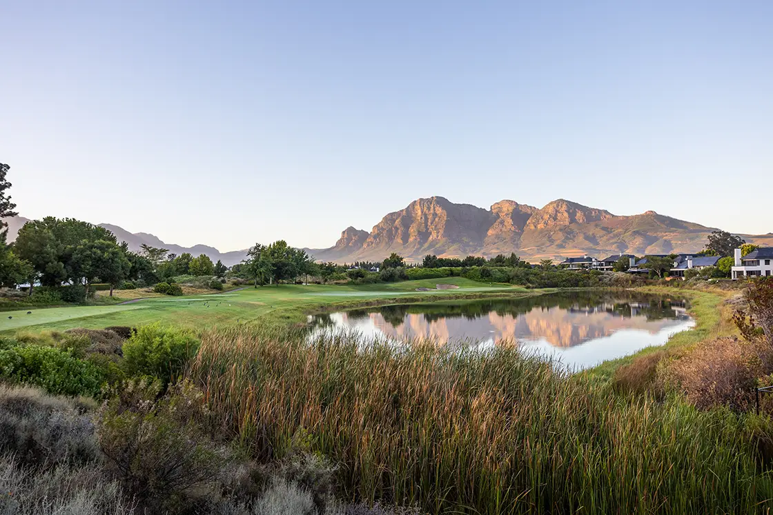 Perfect Hideaways property for sale: A serene landscape of a golf course with a pond reflecting nearby mountains under a clear blue sky. Green vegetation surrounds the tranquil scene. Coral Tree House, Paarl.