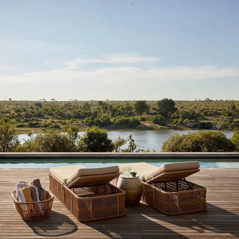 Perfect Hideaways property for sale: A serene poolside view with two wicker loungers overlooking a tranquil river. The scene is framed by lush greenery and blue sky. Ukuthula House in Mpumalanga.