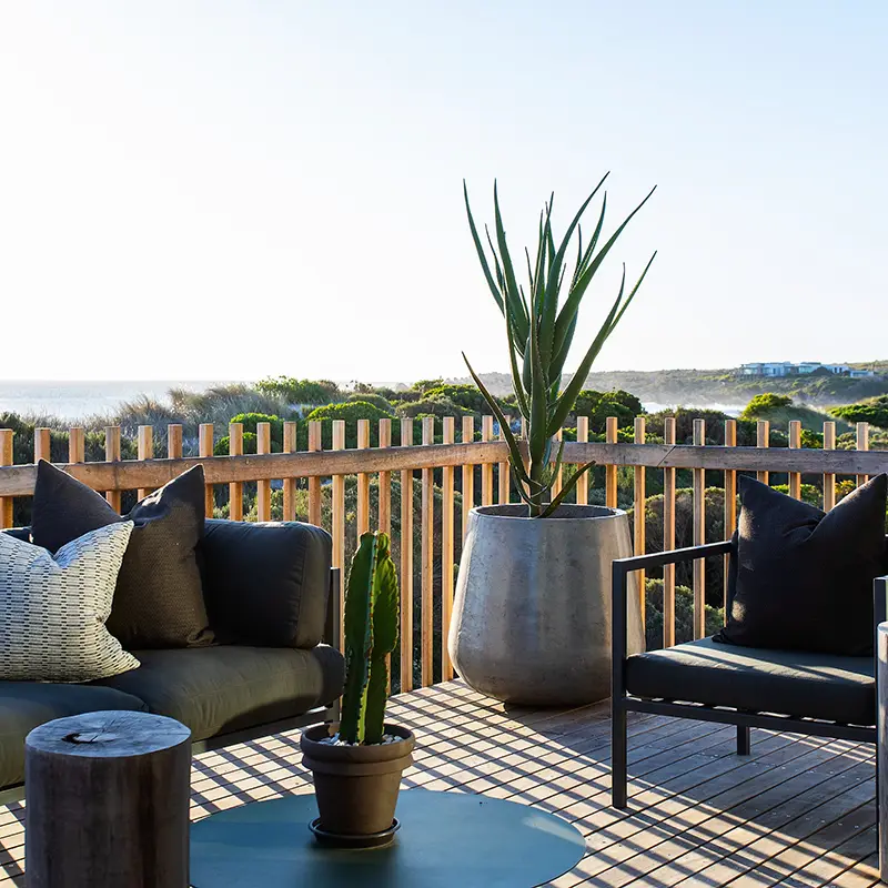 Perfect Hideaways property for sale: Cozy outdoor patio with black cushioned seating, wooden accents, and potted plants. Overlooks scenic, sunny landscape with distant hillside. House On The Dune, Pringle Bay.
