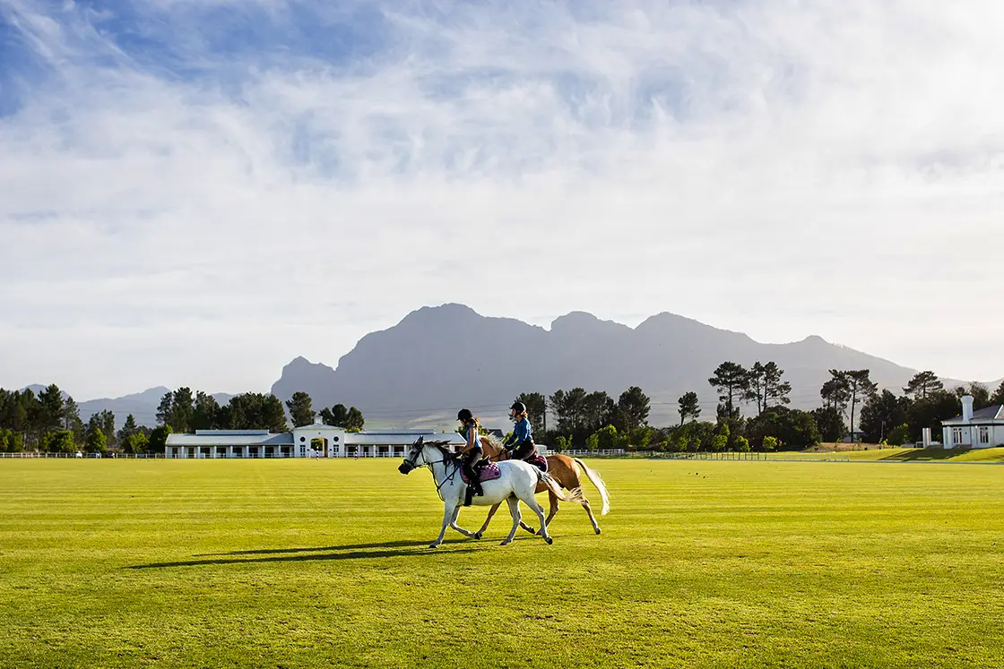 Perfect Hideaways property for sale: Two people riding horses on a lush green field with distant mountains and a clear blue sky in the background. Coral Tree House, Paarl.