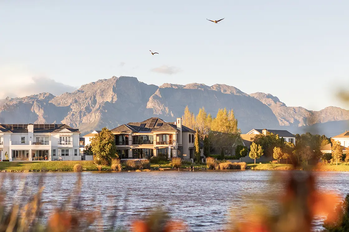 Perfect Hideaways property for sale: Elegant lakeside homes set against a backdrop of majestic mountains at sunset. Birds soar in a clear sky, creating a serene and picturesque scene. Coral Tree House, Paarl.