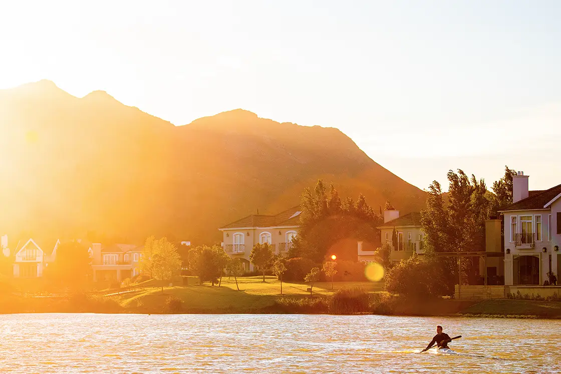 Perfect Hideaways property for sale: A person paddling on a calm river at sunrise, with warm golden light illuminating houses and trees against a backdrop of rolling hills. Coral Tree House, Paarl.