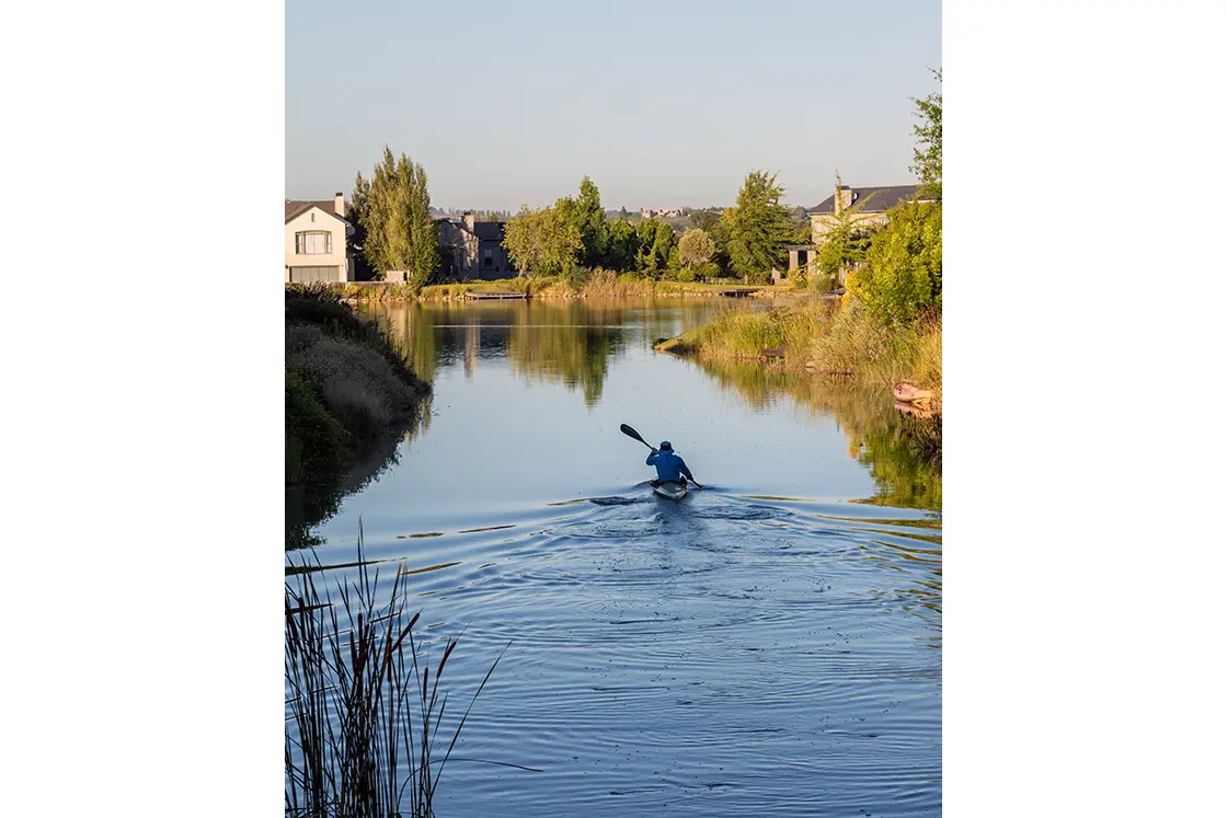 Perfect Hideaways property for sale: A kayaker paddles down a calm river lined with grasses, bordered by houses and trees under a clear blue sky, conveying tranquility and peacefulness. Coral Tree House, Paarl.