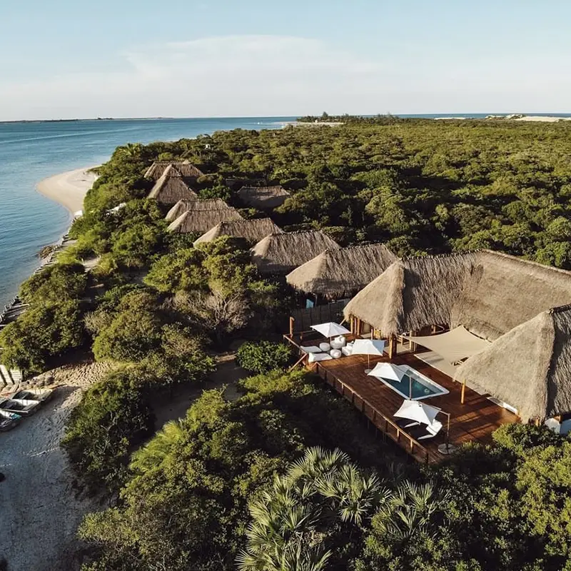 Perfect Hideaways real estate property: Aerial view of a serene beachfront with lush greenery and thatched-roof huts. A wooden deck with umbrellas and a pool overlooks the calm blue ocean. Rio Azul Eco Lodge, Vilanculos.