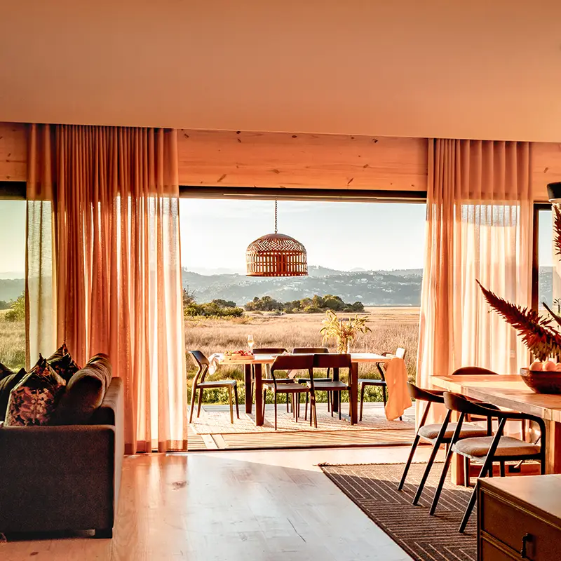 Perfect Hideaways real estate property: Spacious living room with open sliding doors revealing scenic fields and mountains. Warm lighting, soft curtains, and rustic decor create a cozy ambiance. Knysna, Lagoon Song.