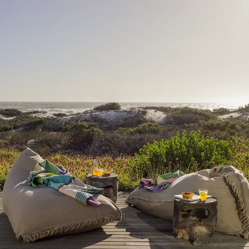 Perfect Hideaways real estate property: Cozy beach scene with two beanbags adorned with colorful throws on a wooden deck, overlooking dunes and ocean. Drinks on rustic tables, calm vibe. Heron Beach House, Jakkalsfontein.