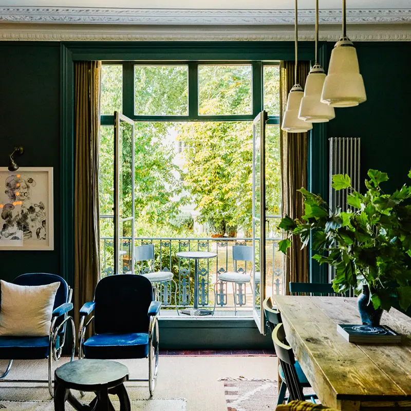 Perfect Hideaways real estate property: Bright living room with open french doors to a small balcony overlooking lush green trees. Blue chairs and a rustic wooden table create a cozy, inviting feel. Alma Square, St John’s Wood.