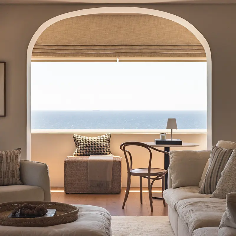 Perfect Hideaways real estate property: Cozy living room with neutral tones, featuring a plush sofa, wicker ottoman, wooden chair, small desk, and stunning ocean view through a large arched window. Lisbon Living Room, Sea Point.