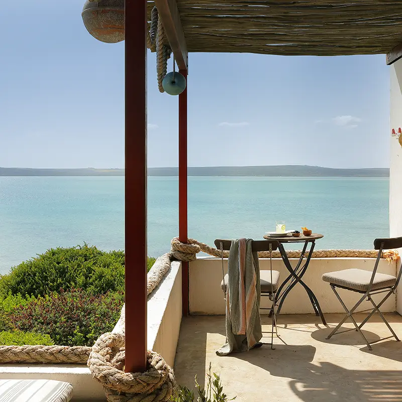 Perfect Hideaways real estate property: Cozy seaside patio with two chairs and a small table under a shaded wooden canopy. Overlooks a tranquil turquoise sea and lush greenery, exuding relaxation. Fynbos Cottage, Churchhaven.