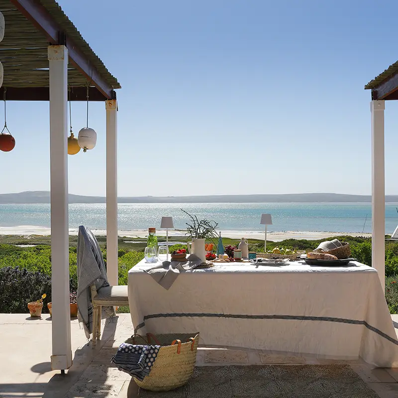 Perfect Hideaways real estate property: Outdoor dining setup under a pergola by a sunny beach. Table with a white cloth, fresh fruits, and drinks. Calm sea in the background, creating a tranquil mood. Lagoon View, Churchhaven.