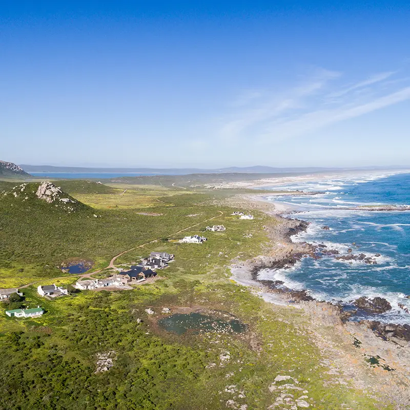 Perfect Hideaways real estate property: Aerial view of a rugged coastline with waves crashing against rocks, green landscape dotted with houses, and a bright blue sky above, conveying tranquility. Kraalhuis, Postberg Nature Reserve.