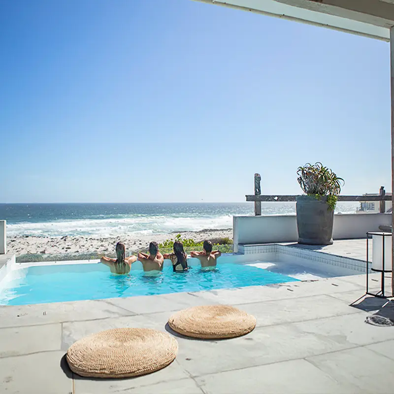 Perfect Hideaways real estate property: Four people relax in a pool by the ocean on a sunny day. The setting is serene, with a blue sky, calm waves, and minimalist decor. Gong Beach House, Yzerfontein.