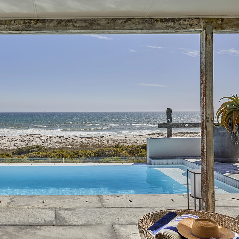 Perfect Hideaways real estate property: View of a serene beachfront with a tranquil infinity pool. A straw hat and book on a table create a relaxed, inviting atmosphere under a clear blue sky. Gong Beach House, Yzerfontein.
