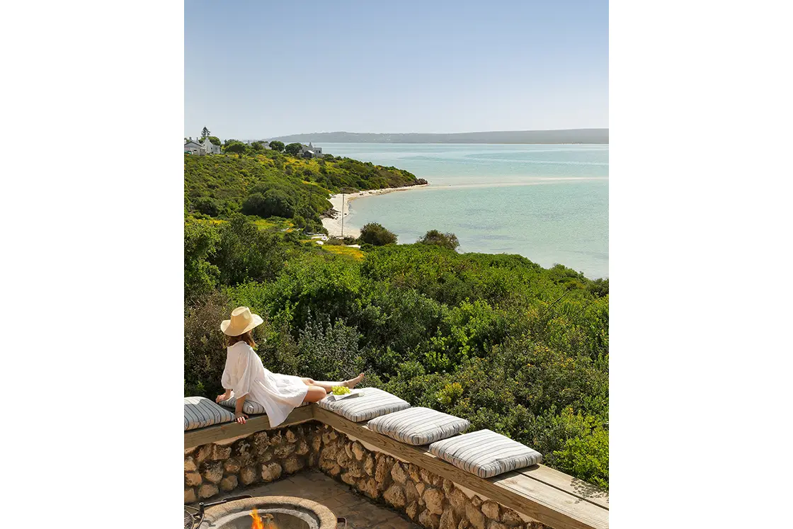 Perfect Hideaways real estate property: A person in a white dress and sun hat lounges on a stone patio bench with cushions, overlooking lush greenery and a tranquil, sunny coastal landscape. Fynbos Cottage, Churchhaven.