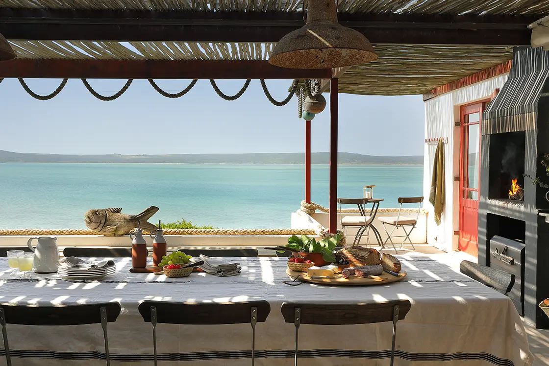 Perfect Hideaways real estate property: Outdoor dining area overlooking a serene blue sea, with a long table set for a meal. Rustic decor, BBQ fire, and coastal ambiance suggest relaxation. Fynbos Cottage, Churchhaven.