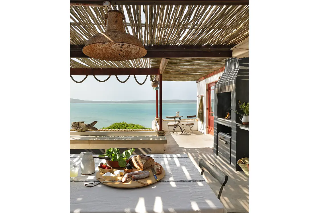 Perfect Hideaways real estate property: Outdoor dining area overlooking the sea, with a table set with bread, cheese, and fruit. A rustic pergola provides shade, creating a serene, inviting atmosphere. Fynbos Cottage, Churchhaven.