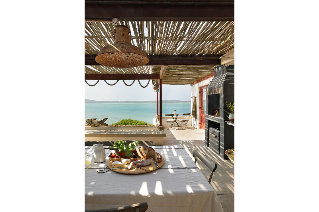 Perfect Hideaways real estate property: Outdoor dining area overlooking the sea, with a table set with bread, cheese, and fruit. A rustic pergola provides shade, creating a serene, inviting atmosphere. Fynbos Cottage, Churchhaven.