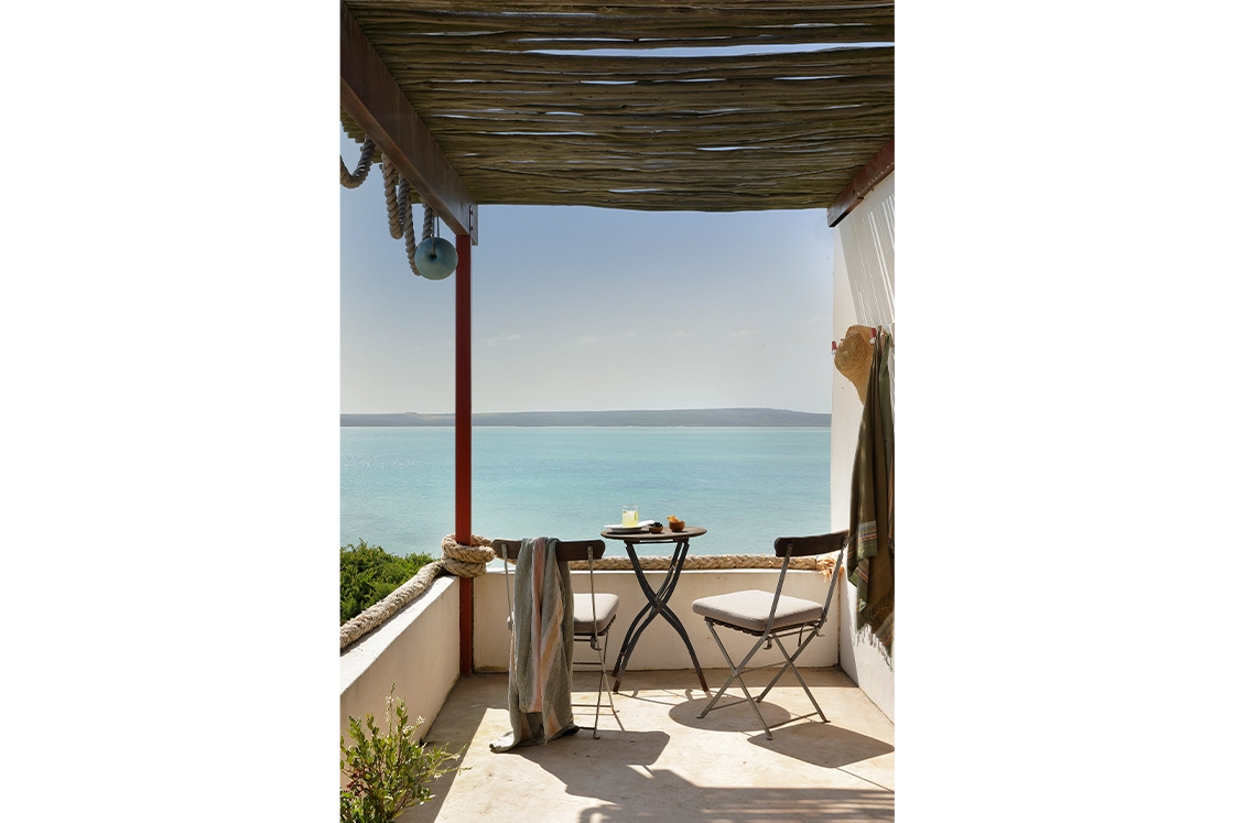 Perfect Hideaways real estate property: Cozy seaside patio with a wooden pergola. Two cushioned chairs and a small table holding drinks overlook a calm, turquoise ocean under a clear sky. Fynbos Cottage, Churchhaven.