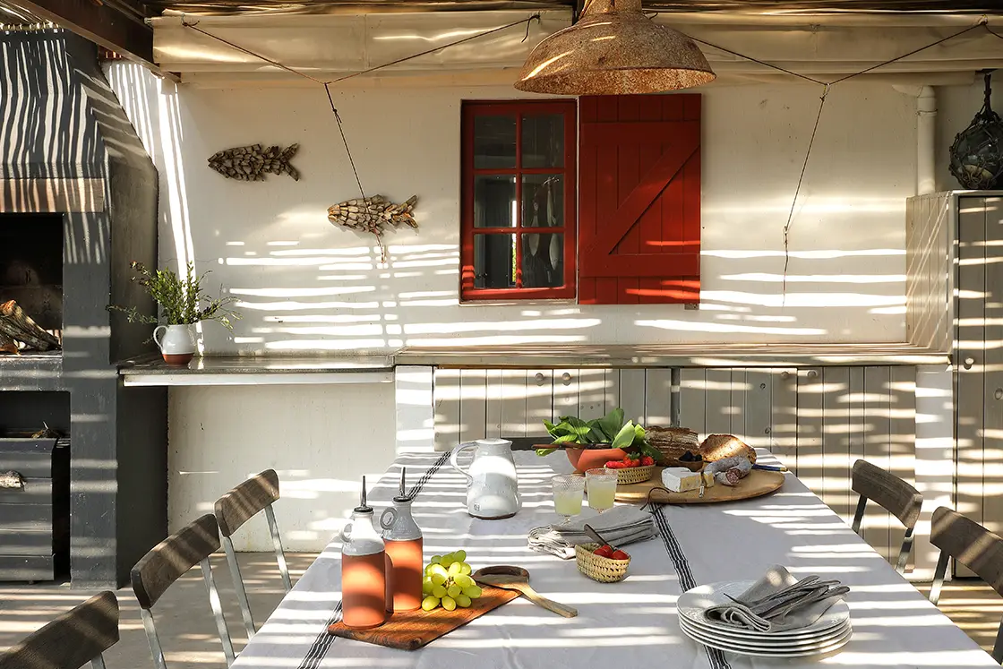 Perfect Hideaways real estate property: Sunlit rustic kitchen with a table set for a meal; bread, grapes, and drinks on a white tablecloth. Warm, inviting atmosphere with shadow patterns. Fynbos Cottage, Churchhaven.