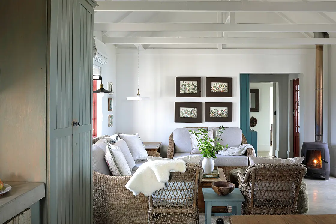 Perfect Hideaways real estate property: Cozy living room with wicker furniture, gray sofa, and a wood-burning stove. Walls decorated with botanical prints, creating a calm, rustic ambiance. Fynbos Cottage, Churchhaven.
