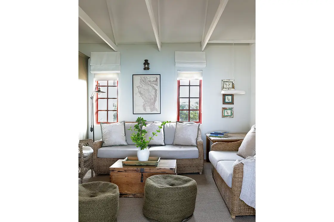 Perfect Hideaways real estate property: Cozy living room with wicker furniture, light gray cushions, a wooden chest table, green poufs, and leafy plant. Two windows with red frames brighten the space. Fynbos Cottage, Churchhaven.