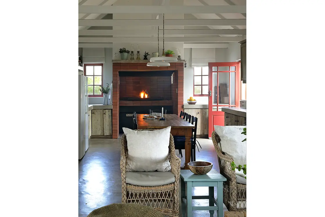 Perfect Hideaways real estate property: Cozy kitchen with a lit fireplace, red-brick chimney, wooden dining table, wicker chairs, red door, and white walls creating a warm, welcoming ambiance. Fynbos Cottage, Churchhaven.