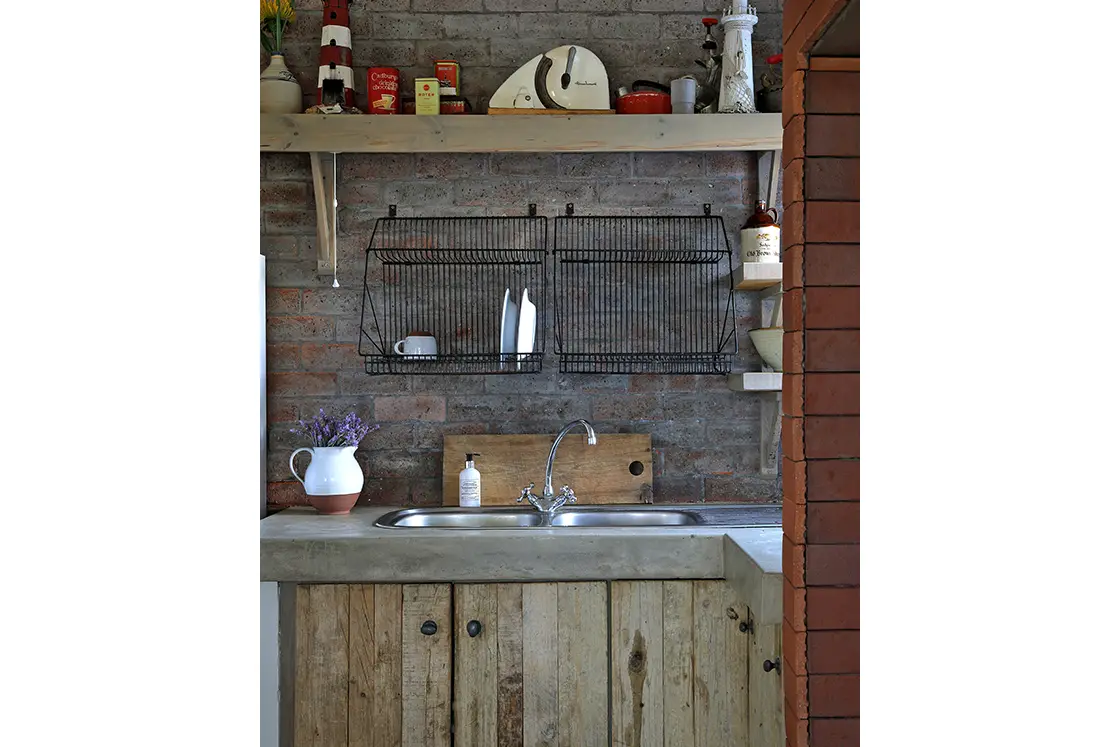 Perfect Hideaways real estate property: Rustic kitchen with a wooden cabinet and concrete countertop. The brick wall features black wire racks and open shelves holding various items. Cozy and vintage feel. Fynbos Cottage, Churchhaven.
