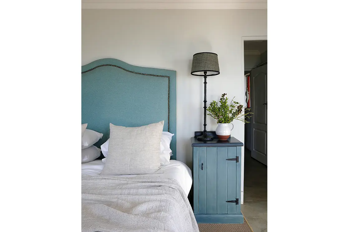 Perfect Hideaways real estate property: Cozy bedroom with a blue headboard, white pillows, and a beige throw. A teal nightstand holds a black lamp and a vase with greenery, creating a calm ambiance. Fynbos Cottage, Churchhaven.