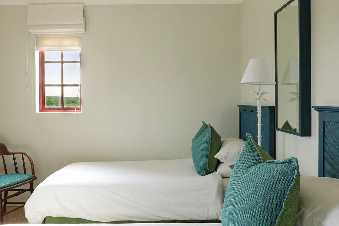 Perfect Hideaways real estate property: Minimalist bedroom with a single bed, white bedding, and teal cushions. A wooden chair near a small window with soft light adds a calming tone. Fynbos Cottage, Churchhaven.