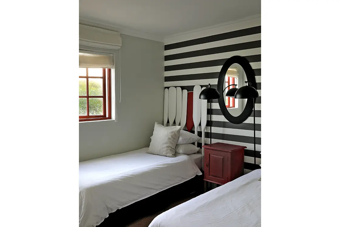 Perfect Hideaways real estate property: Cozy bedroom with two single beds, white linens, oar-themed wall decor, black and white striped wallpaper, red nightstand, and window with soft light. Fynbos Cottage, Churchhaven.
