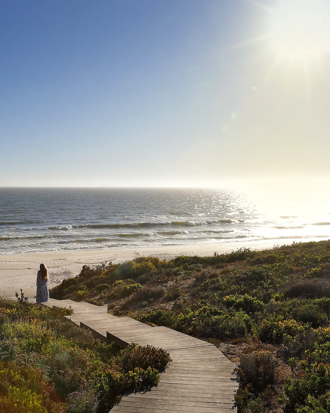 Perfect Hideaways real estate property: A lone woman stands on a wooden path leading to a sunny beach. The ocean sparkles under the bright sun, surrounded by lush greenery, evoking tranquility.