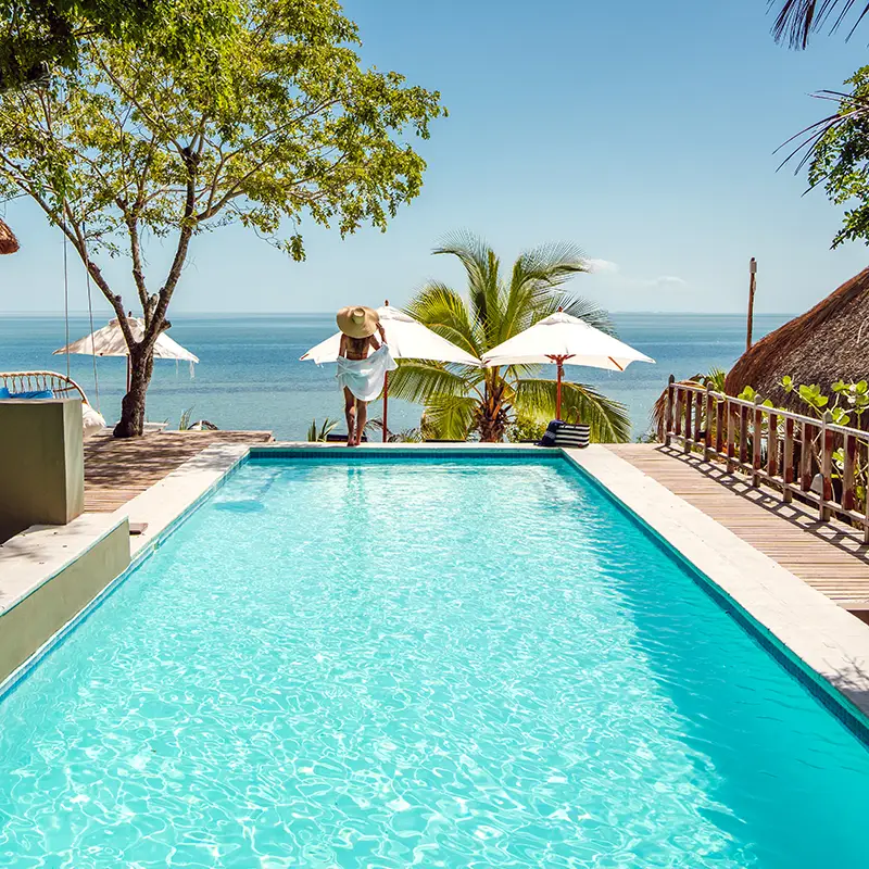Perfect Hideaways real estate property: A serene beachfront view with a sparkling pool in the foreground. A woman in a sun hat stands near the pool edge, overlooking the ocean and palm trees in a relaxed, sunny setting. Mozawi Lodge, The Sanctuary Private Reserve.