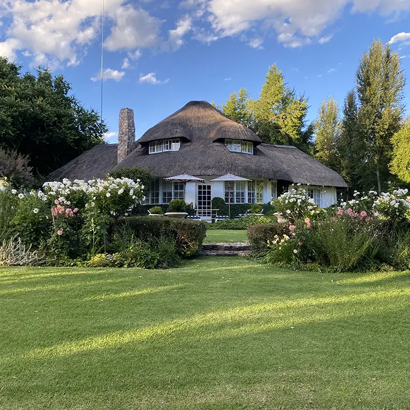 Perfect Hideaways real estate property: Charming house with a thatched roof surrounded by lush gardens and blooming flowers, set against a backdrop of clear blue sky and fluffy clouds. Shangri-La, Vanderbijlpark.