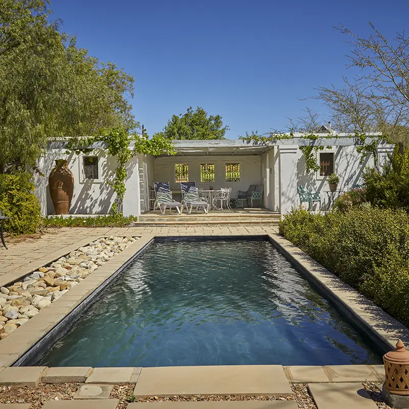 Perfect Hideaways real estate property: A serene backyard features a rectangular pool surrounded by stone tiles, a small rock garden, and lush greenery. A white pergola with lounge chairs is in the background, set against a clear blue sky. Chalcedony Guesthouse, McGregor.