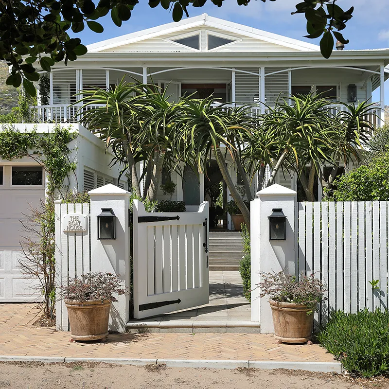 Perfect Hideaways real estate property: White coastal house with a gabled roof, tropical plants, and open gate. The fence and gate evoke a welcoming, serene atmosphere under blue skies. The Boat House, Scarborough.
