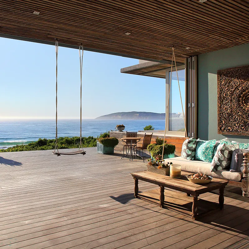 Perfect Hideaways real estate property: Spacious beachside patio with wooden flooring features a swing, cozy seating with turquoise cushions, a table, and ocean view under a clear blue sky. Beachy Head House, Plettenberg Bay.