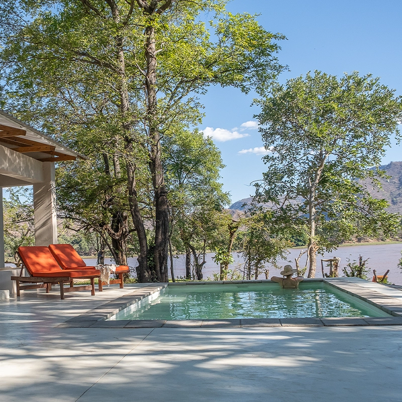 Perfect Hideaways real estate property: A serene poolside scene with wooden deck chairs and orange cushions by a lush green forest, overlooking a tranquil lake under a clear blue sky. Rufunsa game management area, Sendeka River Camp.