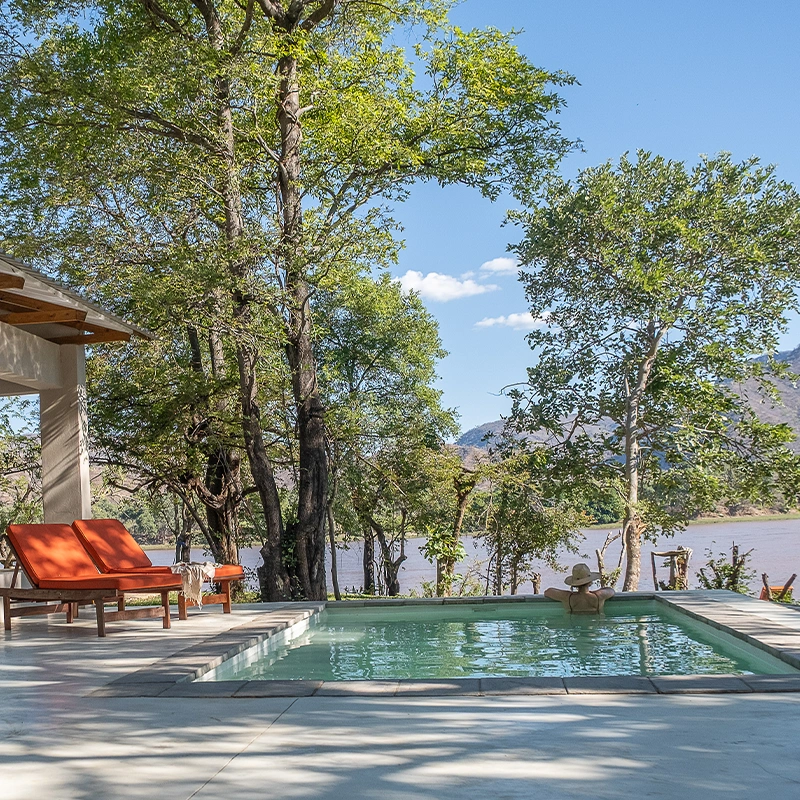 Perfect Hideaways real estate property: A serene poolside scene with wooden deck chairs and orange cushions by a lush green forest, overlooking a tranquil lake under a clear blue sky. Rufunsa game management area, Sendeka River Camp.