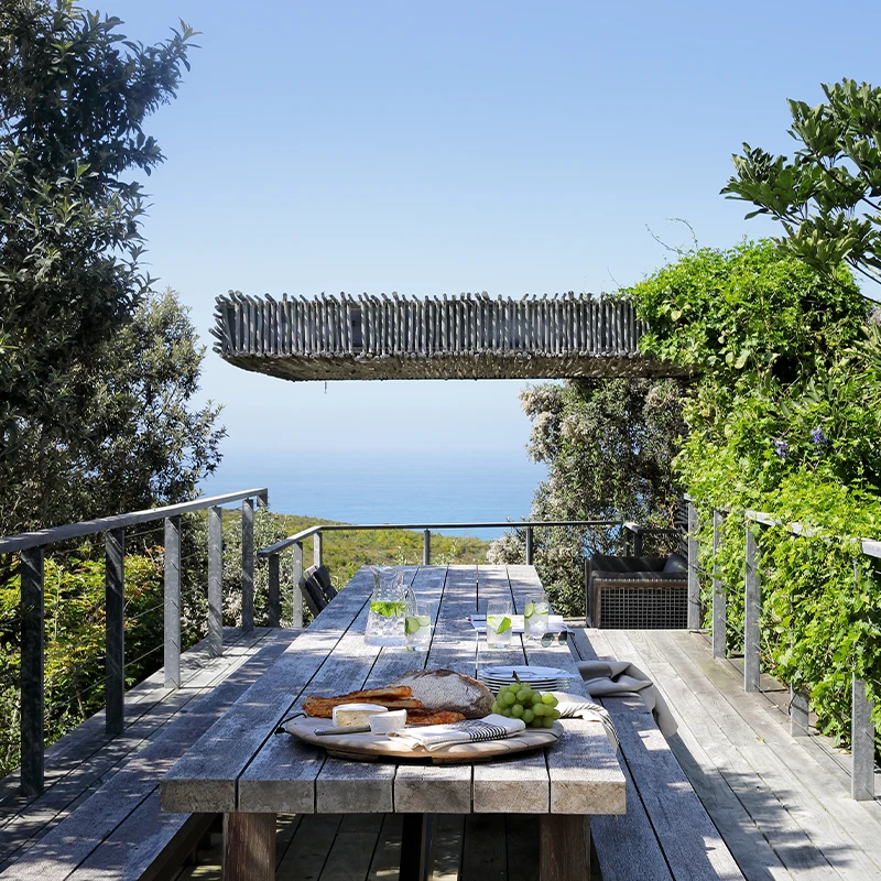 Perfect Hideaways real estate property: Rustic outdoor dining area with wooden table set for lunch, featuring bread and cheese. Surrounded by lush greenery, overlooking a serene ocean view. Ocean's Edge, Knysna.