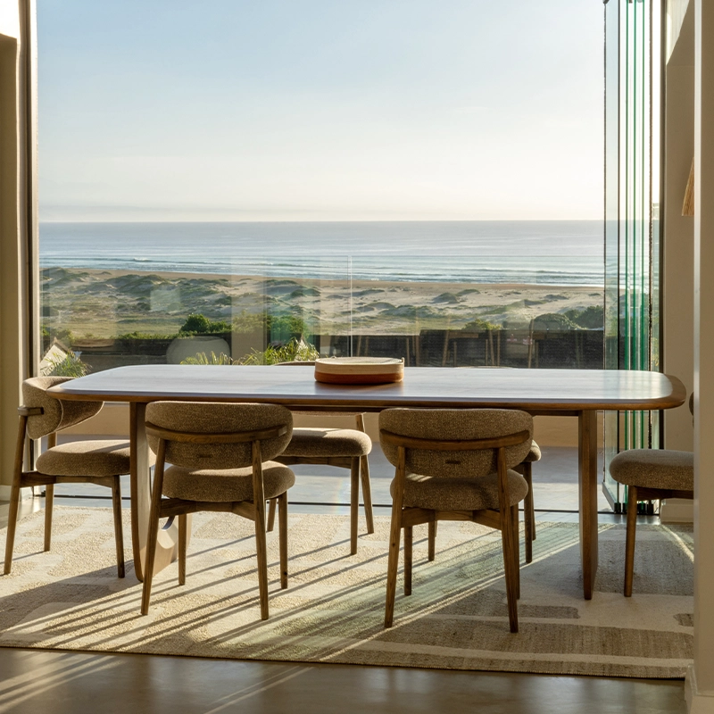 Perfect Hideaways real estate property: Spacious dining room with a wooden table and six chairs facing large windows. The ocean and sandy beach are visible, creating a serene, airy atmosphere. Plettenberg Bay, De Meermin.