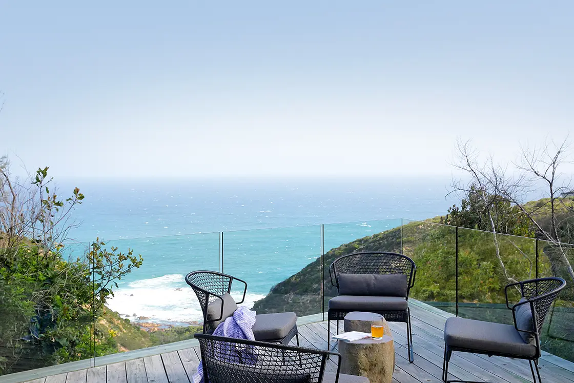 Perfect Hideaways real estate property: A serene coastal deck with modern black wicker chairs overlooking the ocean. Glass railings enhance the panoramic view, suggesting relaxation and tranquility. Knysna, Ocean’s Edge.