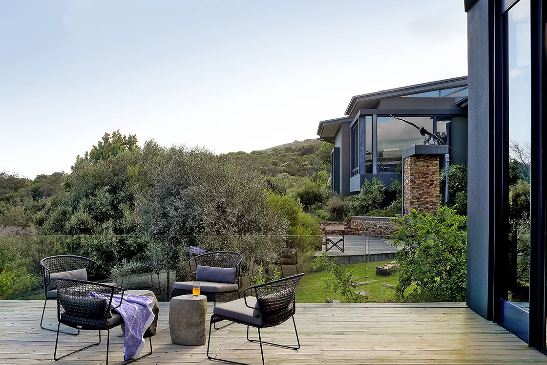 Perfect Hideaways real estate property: Modern patio with black chairs and a small table on a wooden deck. Overlooks lush greenery and a contemporary house with large windows. Calm and inviting atmosphere. Knysna, Ocean’s Edge.