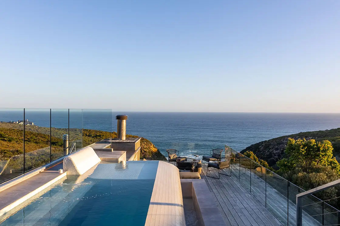 Perfect Hideaways real estate property: A luxurious infinity pool overlooking a vast ocean view, surrounded by glass railings. Adjacent is a wooden deck with lounge chairs, conveying tranquility. Knysna, Ocean’s Edge.