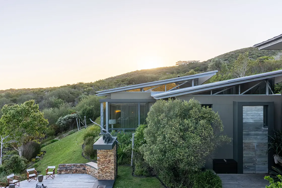 Perfect Hideaways real estate property: Modern hillside house at sunset, featuring slanted roofs, large windows, and a lush garden. Wooden deck with chairs offers a serene, natural setting. Knysna, Ocean’s Edge.