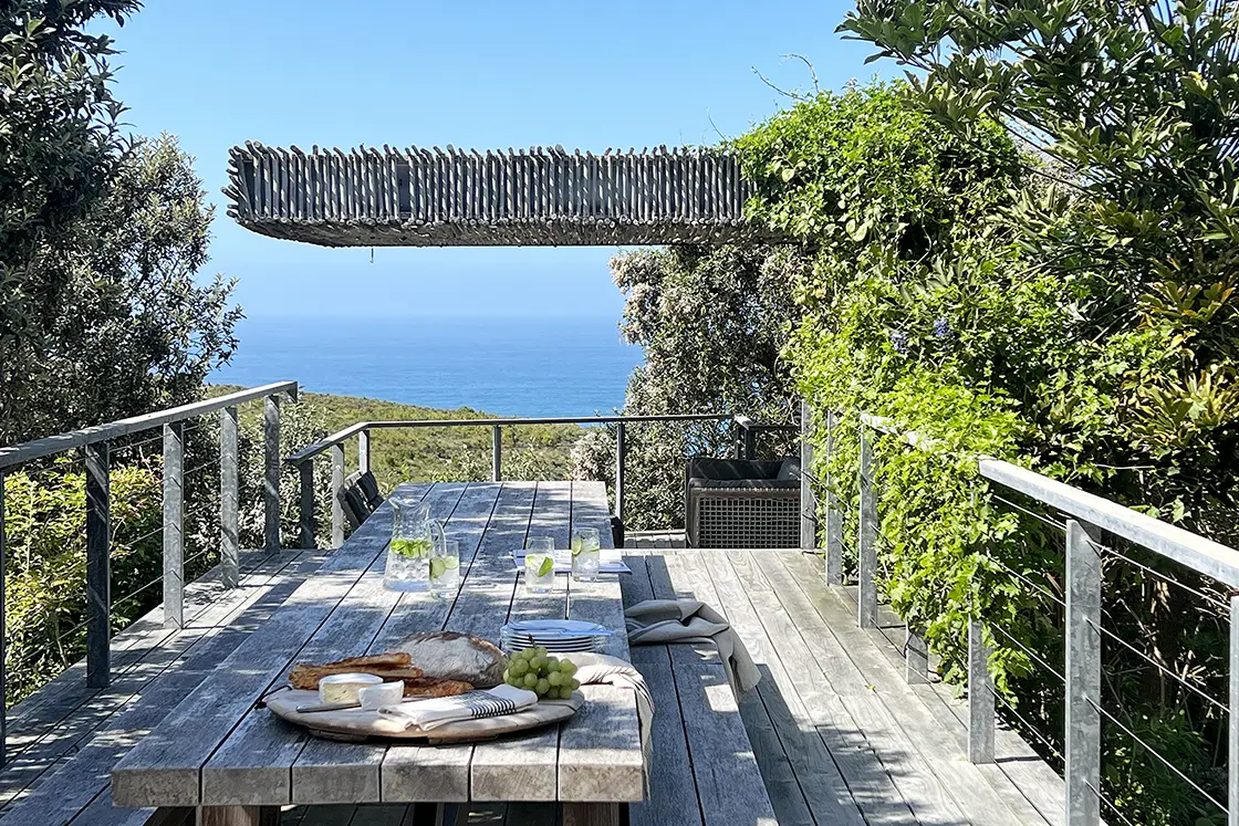 Perfect Hideaways real estate property: A rustic wooden table with bread, grapes, and drinks overlooks a serene ocean view. Surrounded by lush greenery, the setting feels peaceful and inviting. Knysna, Ocean’s Edge.